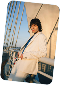 Man in a white suit leaning on a bridge railing with cables in the background at sunset.